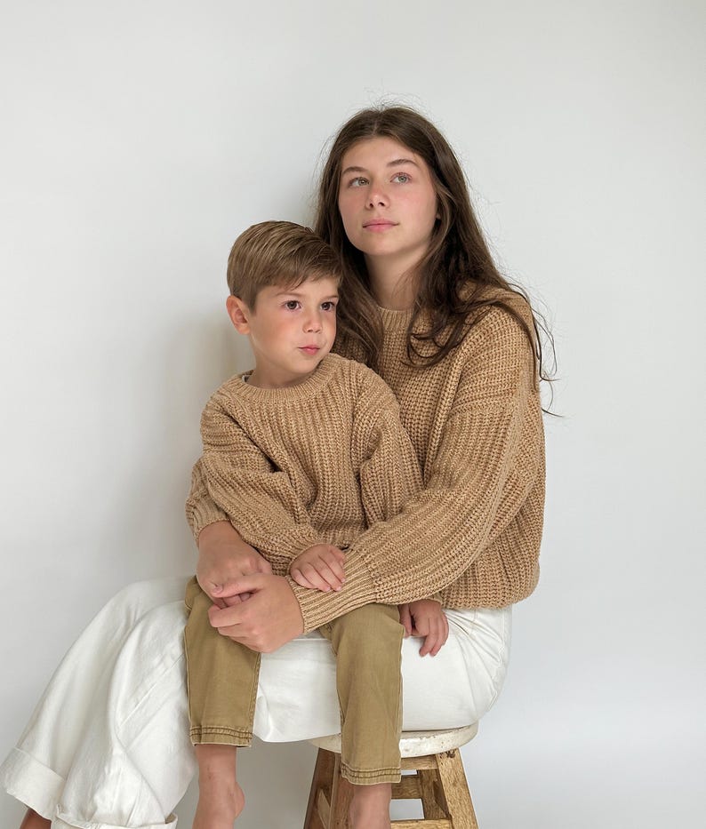 May include: Two matching tan long-sleeve sweaters with a thick ribbed knit texture and crew necklines. The sweaters feature a chunky weave pattern, long sleeves with ribbed cuffs, and a relaxed fit, providing a uniform look for two individuals.