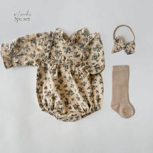 May include: A 3-piece baby outfit set featuring a long-sleeved floral print romper, a headband with a bow, and a pair of beige socks. The text "w/socks 3pc set" is printed on the image.