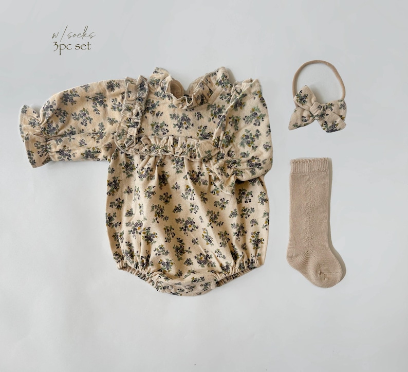 May include: A 3-piece baby outfit set featuring a long-sleeved floral print romper, a headband with a bow, and a pair of beige socks. The text "w/socks 3pc set" is printed on the image.
