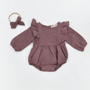 Könnte beinhalten: Ein mauvefarbener Baby-Strampler aus Leinen mit Rüschenärmeln und einem Schleifen-Haarband. Der Strampler hat einen runden Ausschnitt und Druckknöpfe am unteren Ende.