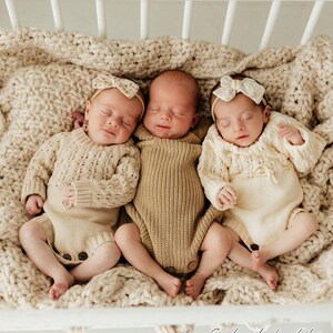 May include: Three newborn babies wearing knitted outfits and headbands with floral patterns are sleeping in a white crib. The crib is lined with a cream-colored knitted blanket.