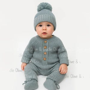 May include: A baby wearing a matching knit set in a muted teal color. The set includes a beanie with a pom-pom, a button-down romper, and booties. The romper has wooden buttons. The baby is sitting on a white background.