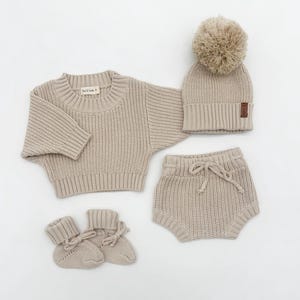 Peut inclure: Un ensemble de vêtements en tricot beige pour bébé comprenant un pull, un short, un bonnet et des chaussons. Le bonnet est surmonté d'un pompon moelleux.