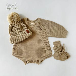 May include: A 3-piece baby outfit set in a light brown color. The set includes a knitted sweater bodysuit, a knitted hat with a pom-pom, and knitted booties. The outfit is made of soft, comfortable material and is perfect for keeping baby warm and cozy.