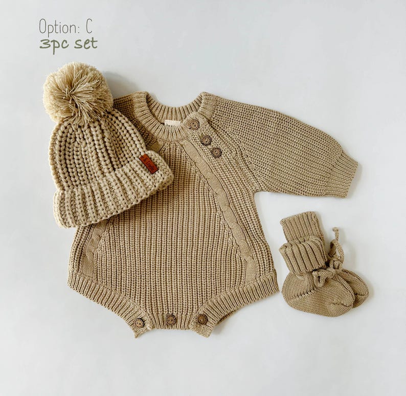 May include: A 3-piece baby outfit set in a light brown color. The set includes a knitted sweater bodysuit, a knitted hat with a pom-pom, and knitted booties. The outfit is made of soft, comfortable material and is perfect for keeping baby warm and cozy.