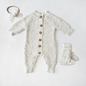 May include: A white knitted baby onesie with brown buttons, a matching headband, and booties. The onesie has a speckled pattern.