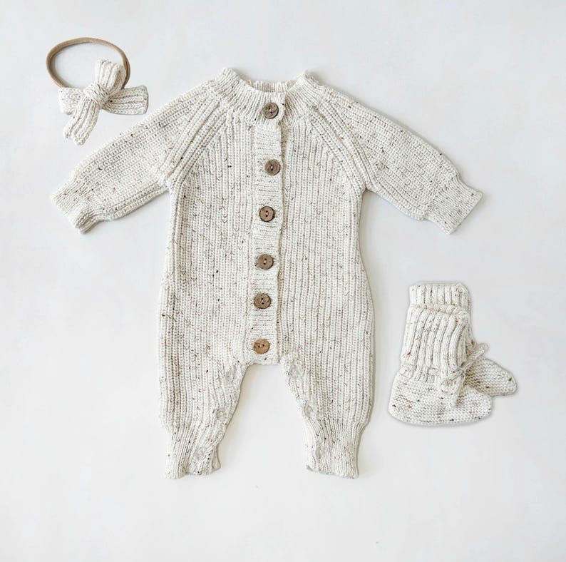 May include: A white knitted baby onesie with brown buttons, a matching headband, and booties. The onesie has a speckled pattern.