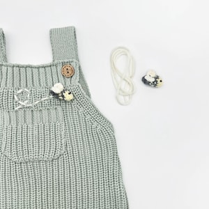 May include: A light gray knitted baby overall with wooden buttons and a front pocket. The overall is decorated with a small crocheted bee and a white string. A separate crocheted bee and string are also present.