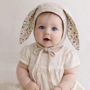 May include: A baby wearing a beige linen bonnet with long, floppy bunny ears. The inside of the ears features a floral pattern with red and pink flowers. The bonnet ties under the chin with a matching beige ribbon. The baby is wearing a beige dress.
