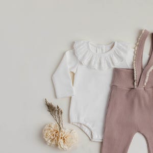 Peut inclure: Un body blanc à manches longues avec un col en dentelle et un pantalon côtelé mauve. Le body a une fermeture à pression à l'entrejambe. Le pantalon a des bretelles réglables.