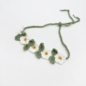 May include: A crocheted floral headband featuring white daisy-like flowers with yellow centers and green leaves. The headband is connected by a green crocheted cord, perfect for adding a touch of nature-inspired charm to any hairstyle.