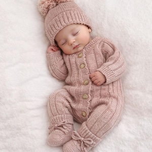 May include: A sleeping baby wearing a knitted pink outfit, including a hat with a pom-pom, a button-down jumpsuit, and matching booties with bows. The outfit is made of a ribbed knit fabric. The baby is lying on a white, fluffy surface.