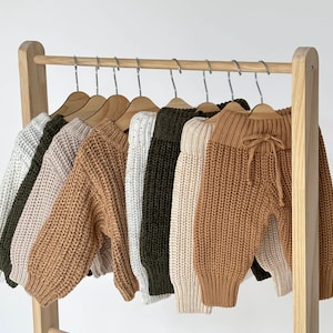 May include: A set of five knitted sweaters and pants in various colors, including beige, brown, white, and green, hanging on a wooden rack.