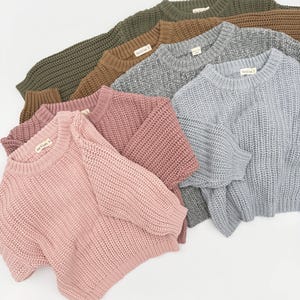 May include: A collection of five long-sleeve knit sweaters in olive green, brown, heather gray, dusty rose, and light blue. Each sweater features a crew neckline, ribbed cuffs, and a hem with a small white tag labeled "Bear & Cradle".