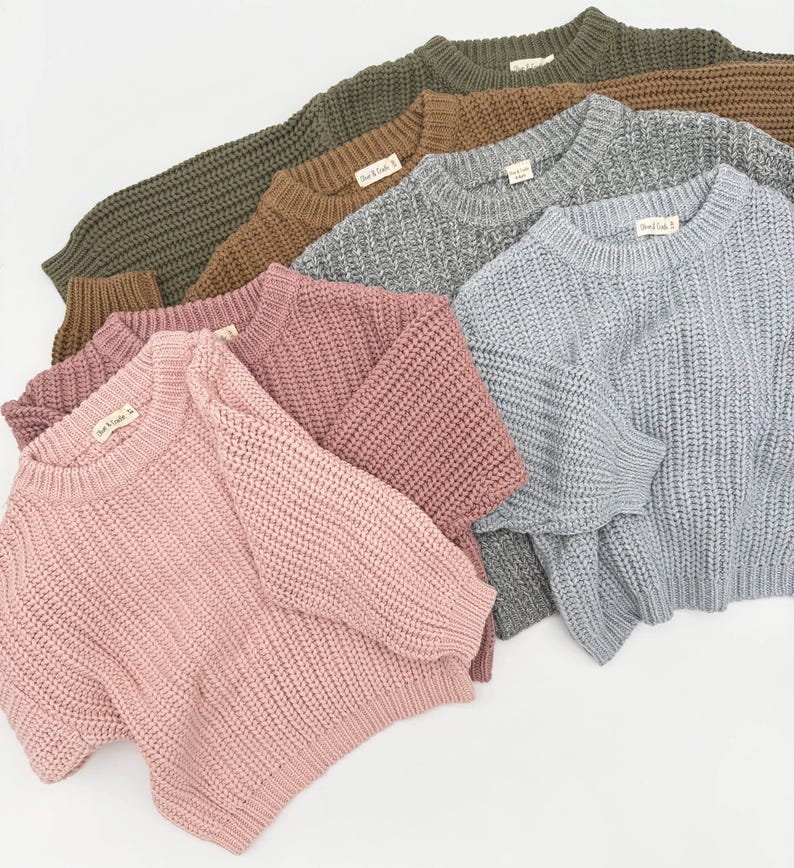 May include: A collection of five long-sleeve knit sweaters in olive green, brown, heather gray, dusty rose, and light blue. Each sweater features a crew neckline, ribbed cuffs, and a hem with a small white tag labeled "Bear & Cradle".