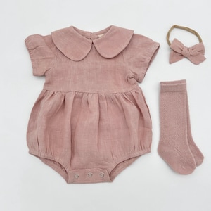 Op de afbeelding: Een stofroze babyromper met een Peter Pan-kraag, korte mouwen en een geplooid taille. Een bijpassende strik-hoofdband en kniehoge sokken maken de set compleet. De romper heeft drukknoopsluitingen.