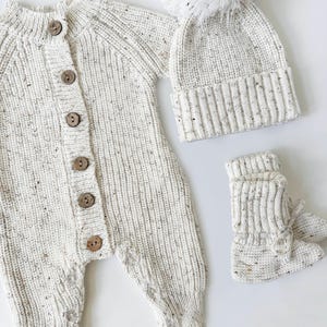 May include: A white knitted baby outfit with a matching hat and booties. The hat has a white pom-pom on top. The outfit has a button closure.