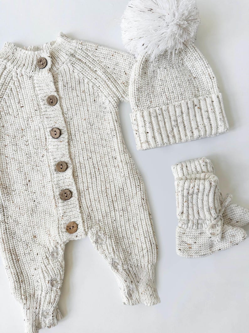 May include: A white knitted baby outfit with a matching hat and booties. The hat has a white pom-pom on top. The outfit has a button closure.