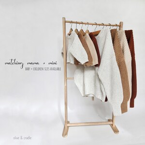 May include: A wooden clothing rack displaying several chunky knit sweaters in cream, tan, and rust colors. The text on the image reads: matching mama + mini BABY + CHILDREN SIZES AVAILABLE and olive & cradle. The sweaters feature long sleeves.