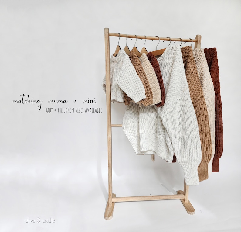 May include: A wooden clothing rack displaying several chunky knit sweaters in cream, tan, and rust colors. The text on the image reads: matching mama + mini BABY + CHILDREN SIZES AVAILABLE and olive & cradle. The sweaters feature long sleeves.