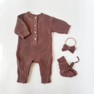 Puede incluir: Un mono de punto color malva para bebé con cierre de botones, una diadema a juego con lazo y un par de botines de punto.