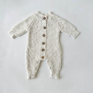 May include: A white knitted baby onesie with brown button closures. The onesie has a speckled pattern.