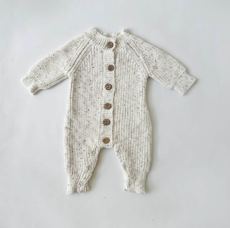 May include: A white knitted baby onesie with brown button closures. The onesie has a speckled pattern.