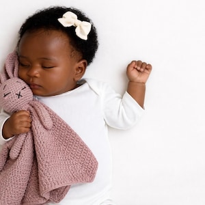 May include: A sleeping baby wearing a white onesie and a cream-colored bow in their hair. The baby is holding a pink crocheted bunny lovey. The lovey has a textured pattern and stitched facial features.