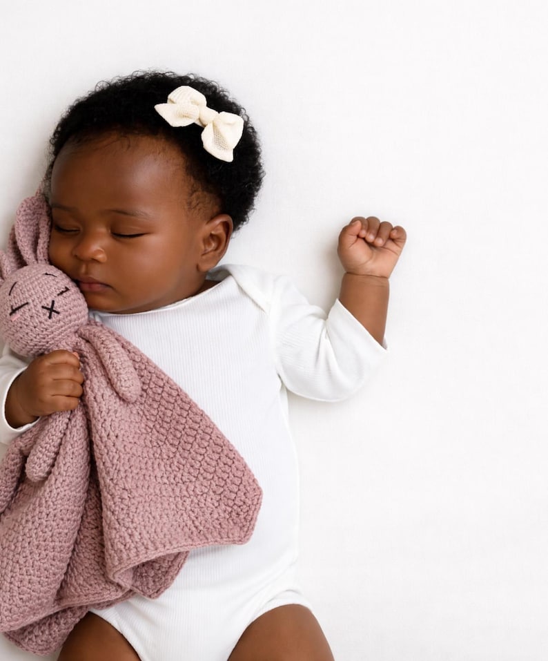 May include: A sleeping baby wearing a white onesie and a cream-colored bow in their hair. The baby is holding a pink crocheted bunny lovey. The lovey has a textured pattern and stitched facial features.