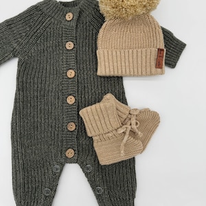 May include: A knitted baby outfit set in a neutral colour palette. The set includes a dark grey romper with wooden buttons, a tan beanie with a large pom-pom, and matching booties. The romper has button closures at the legs.