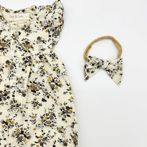 May include: A white baby onesie with a floral print, button closure and ruffled sleeves. The onesie is paired with a matching brown floral headband.