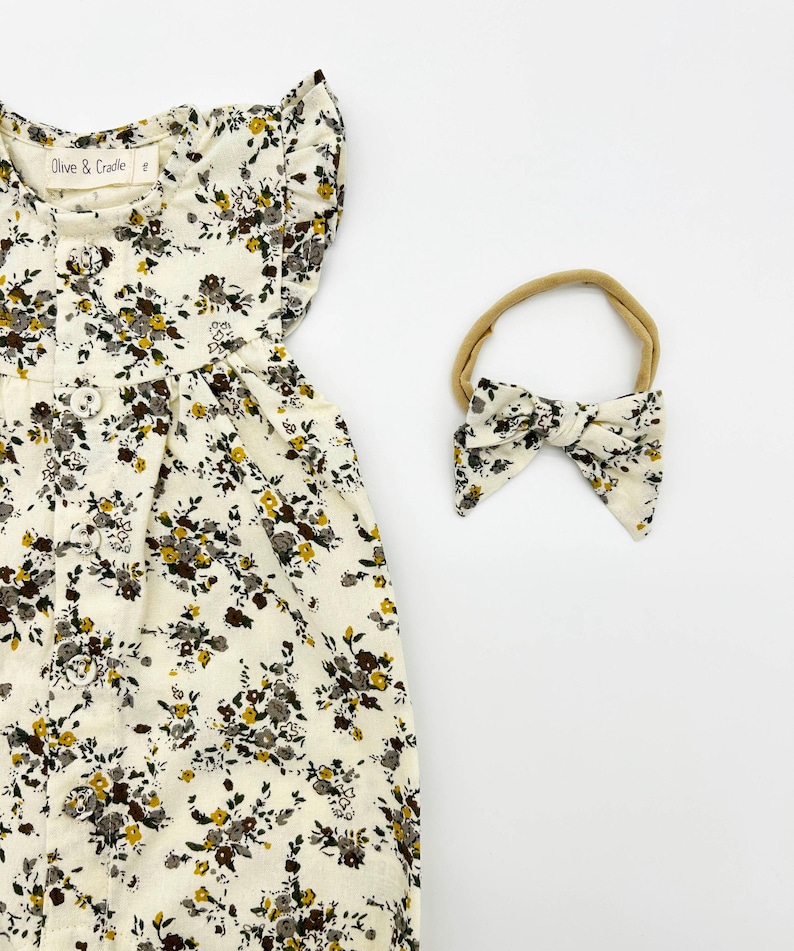 May include: A white baby onesie with a floral print, button closure and ruffled sleeves. The onesie is paired with a matching brown floral headband.