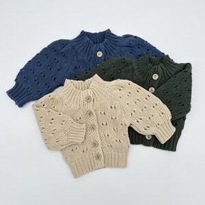 May include: Three knitted cardigans in navy blue, olive green, and beige. Each cardigan has a round collar, wooden buttons, and a textured knit pattern. The sleeves are long and gathered at the wrist. The cardigans are displayed on a white surface.