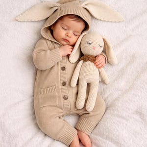 Puede incluir: Un bebé dormido con un traje de conejito de punto beige con orejas grandes y un juguete de conejito de punto a juego. El bebé está acostado sobre una superficie blanca texturizada. El traje de conejito tiene una parte delantera abotonada y un bolsillo.