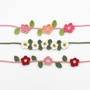 May include: Three rows of crocheted floral garlands. The top row features pink and peach flowers with green leaves on a pink string. The middle row has white flowers with yellow centers and green leaves on a green string. The bottom row has red and pink flowers with green leaves on a pink string.