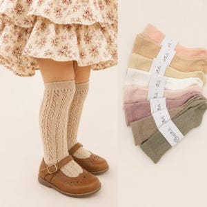 Peut inclure: Une enfant porte des chaussettes hautes beiges à motifs ajourés et des chaussures Mary Jane en cuir marron. Une pile de chaussettes pastel, roses, beiges, blanches et vertes, est présentée. Les chaussettes sont liées par une étiquette.