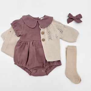 May include: A baby outfit set with a mauve linen romper, a cream knitted cardigan with a lace pattern, and a pair of cream knitted socks. The set also includes a mauve bow.