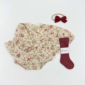 Puede incluir: Un conjunto de ropa de bebé con un pelele con estampado floral con dobladillo con volantes, una diadema con lazo burdeos y calcetines burdeos a juego. El pelele tiene mangas largas y un fondo crema con detalles florales rojos.