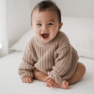 May include: A baby wearing a long-sleeved, chunky knit sweater in a light beige color. The sweater features a ribbed crew neckline, ribbed cuffs at the wrists, and a thick, vertical cable-knit texture throughout the garment's fabric construction.