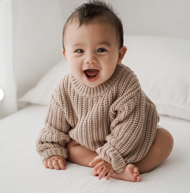 May include: A baby wearing a long-sleeved, chunky knit sweater in a light beige color. The sweater features a ribbed crew neckline, ribbed cuffs at the wrists, and a thick, vertical cable-knit texture throughout the garment's fabric construction.