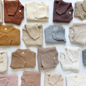 May include: A collection of chunky knit sweaters in various solid colors including cream, rust, mustard, light blue, olive green, grey, and shades of brown. Each sweater features a crew neckline, long sleeves, and a thick, textured ribbed pattern.