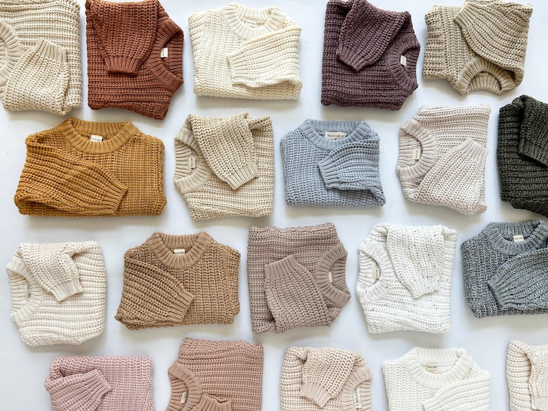May include: A collection of chunky knit sweaters in various solid colors including cream, rust, mustard, light blue, olive green, grey, and shades of brown. Each sweater features a crew neckline, long sleeves, and a thick, textured ribbed pattern.