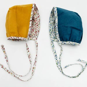 May include: Two baby bonnets with floral trim. One bonnet is mustard yellow and the other is teal blue.