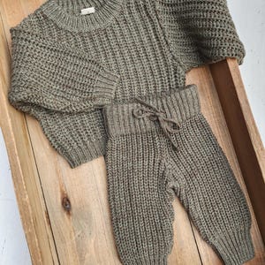 May include: A green knitted sweater and matching pants set for a baby or toddler. The sweater has a round neckline and long sleeves. The pants have a drawstring waistband and a relaxed fit.