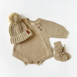 Peut inclure: Un ensemble de vêtements pour bébé en tricot beige avec un bonnet et des chaussons assortis. Le bonnet est surmonté d'un pompon moelleux. La tenue a une fermeture à bouton sur l'épaule et un motif en tricot torsadé.