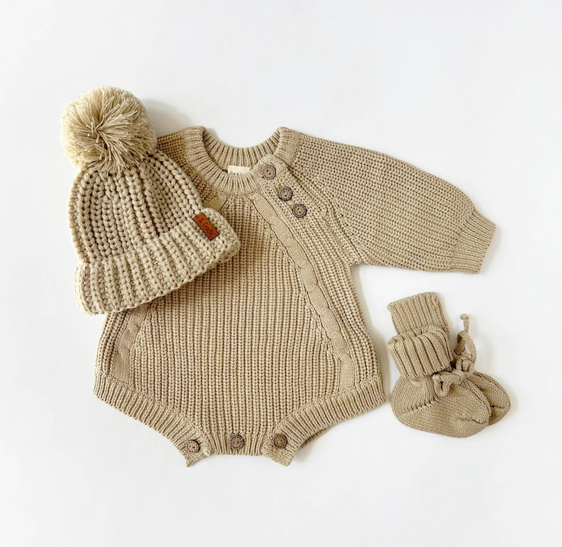 May include: A beige knitted baby outfit set with a matching hat and booties. The hat has a fluffy pom-pom on top. The outfit has a button closure on the shoulder and a cable knit design.