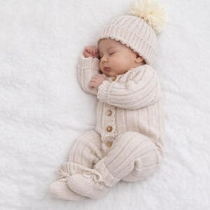 May include: A sleeping baby wearing a cream-colored knitted outfit, including a hat with a pom-pom, a button-down onesie, and matching booties. The outfit is made of a soft, ribbed knit material. The baby is lying on a white, textured surface.