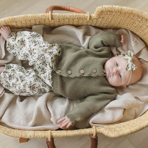 May include: A baby wearing a green cardigan and floral trousers lies in a woven basket. The baby is wearing a floral headband and has a neutral expression.