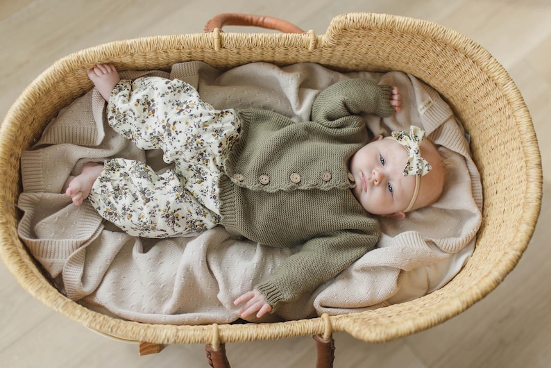 May include: A baby wearing a green cardigan and floral trousers lies in a woven basket. The baby is wearing a floral headband and has a neutral expression.