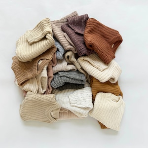 May include: A collection of folded long-sleeve sweaters in various neutral earth tones including cream, beige, tan, brown, grey, and dark purple. Each sweater features a thick, chunky knit waffle texture with a crew neckline and ribbed cuffs.
